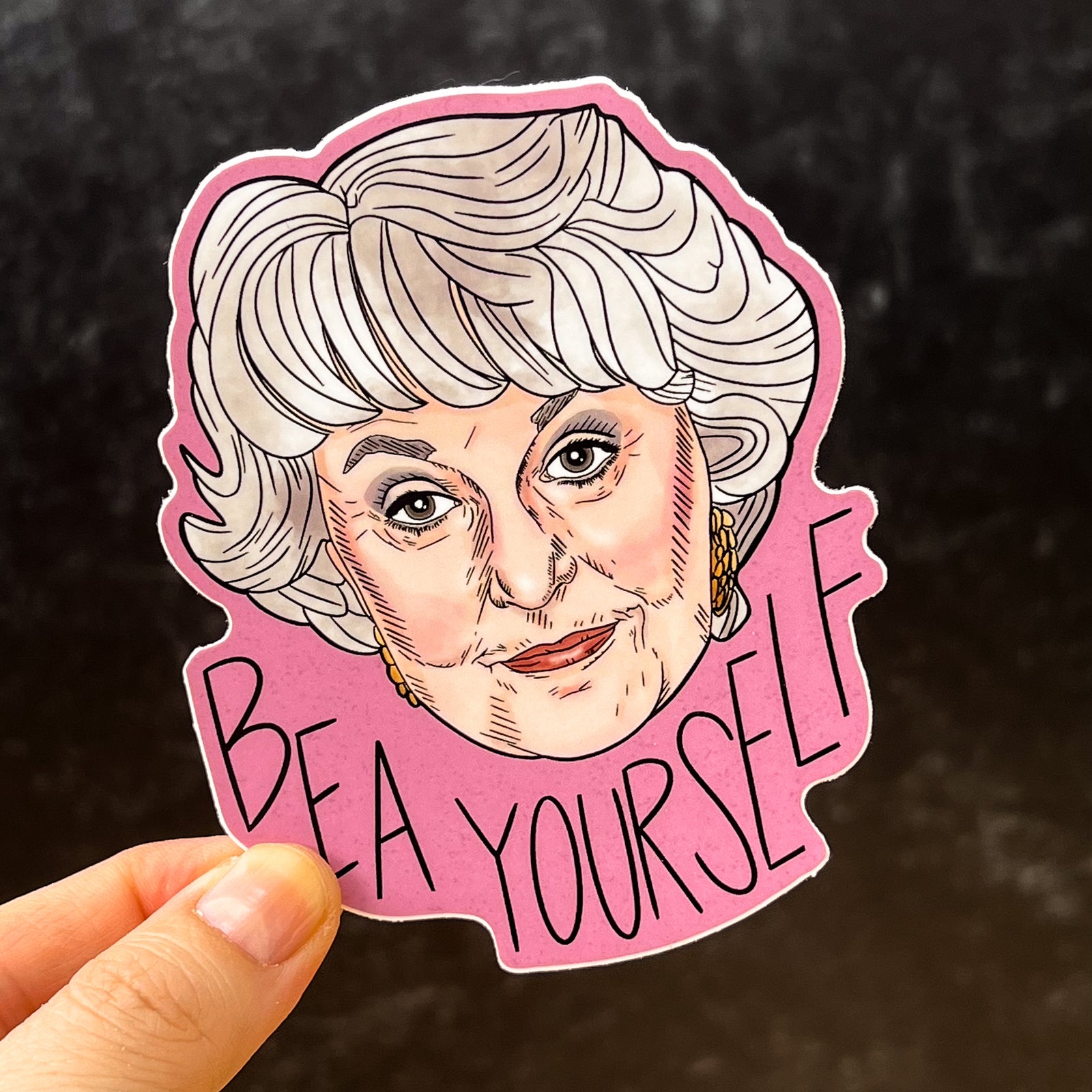 A sticker featuring Bea Arthur and the message "Bea Yourself" is held up against a black background