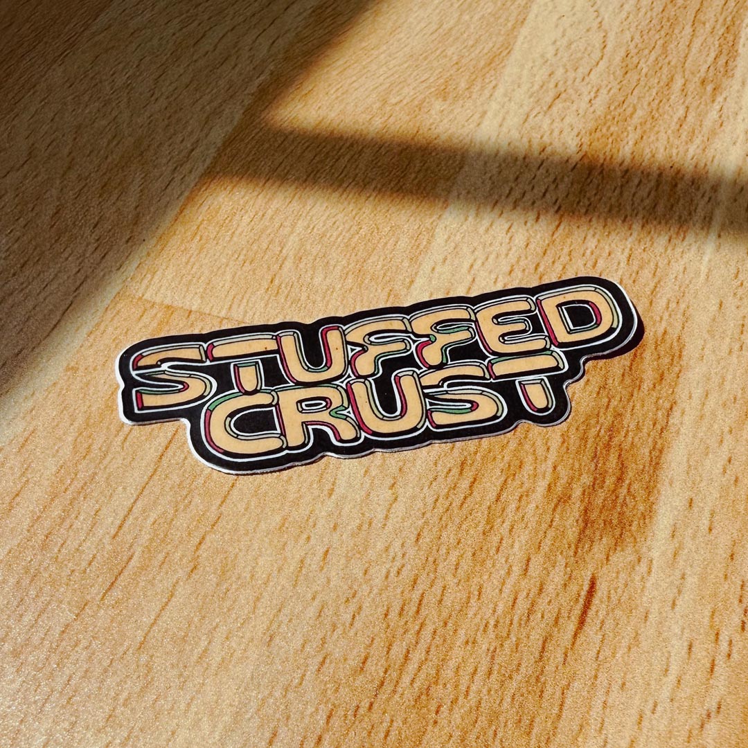 A die-cut sticker reading 'STUFFED CRUST' in a retro bubble font with black outlines and tan lettering, placed on a warm wood surface evocative of places with stuffed crust pizza