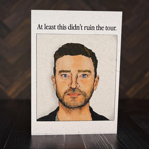 Card with a portrait of a Justin Timberlake and text 'At least this didn't ruin the tour' on a wooden surface.