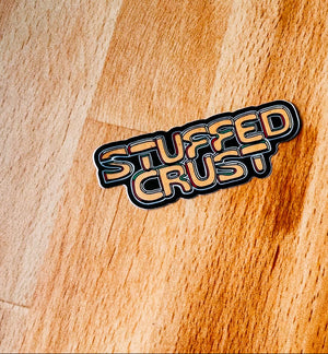 A die-cut sticker reading 'STUFFED CRUST' in a retro bubble font with black outlines and tan lettering, placed on a warm wood surface evocative of places with stuffed crust pizza